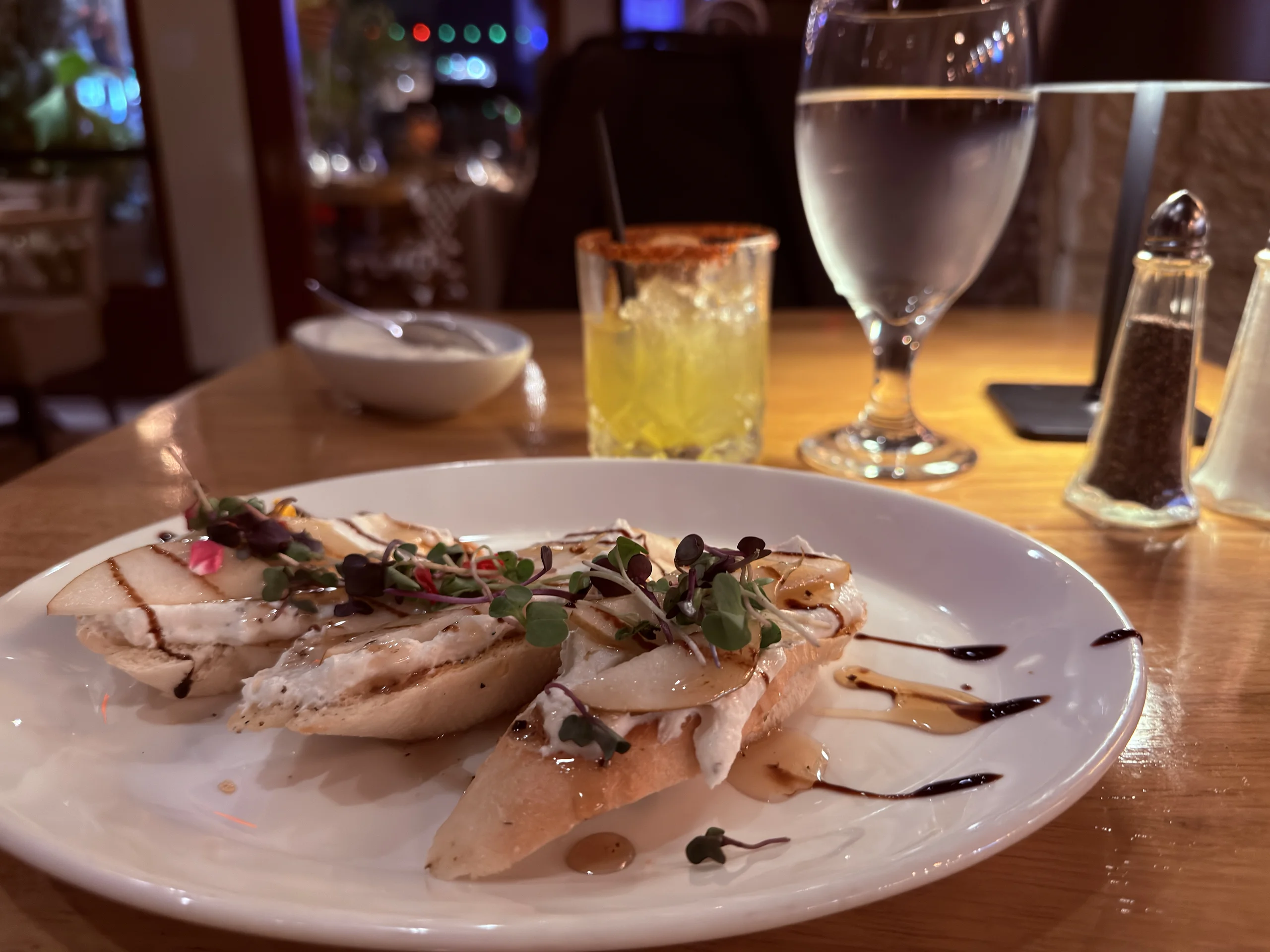 餐桌上的義式烤麵包片,上面鋪著起司與香草,背景有調酒與水杯