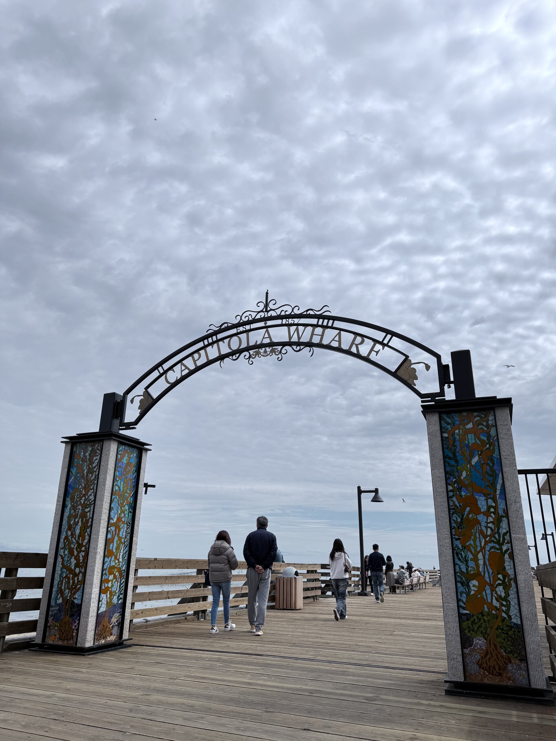 陰天中,幾位遊客漫步在Capitola Wharf木製棧橋上,拱門兩側裝飾有彩繪圖案。