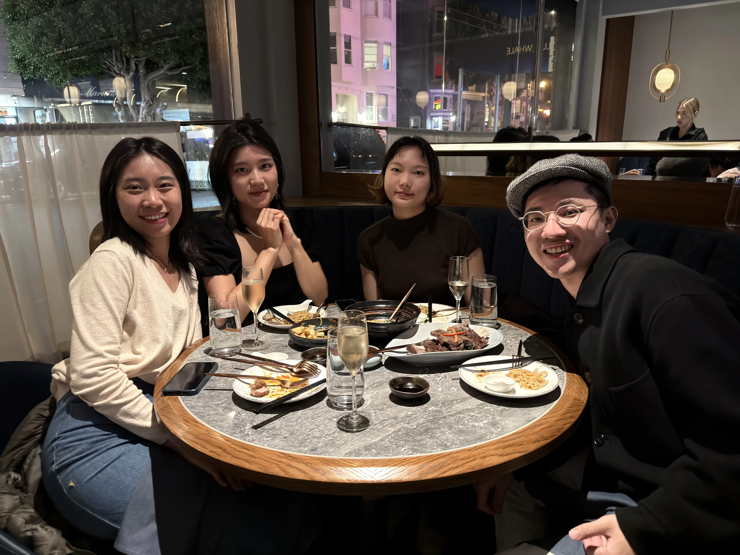 Tomy 與朋友在餐廳圓桌前合影,桌上擺著牛排、香檳與共享餐點,窗外是夜晚街景