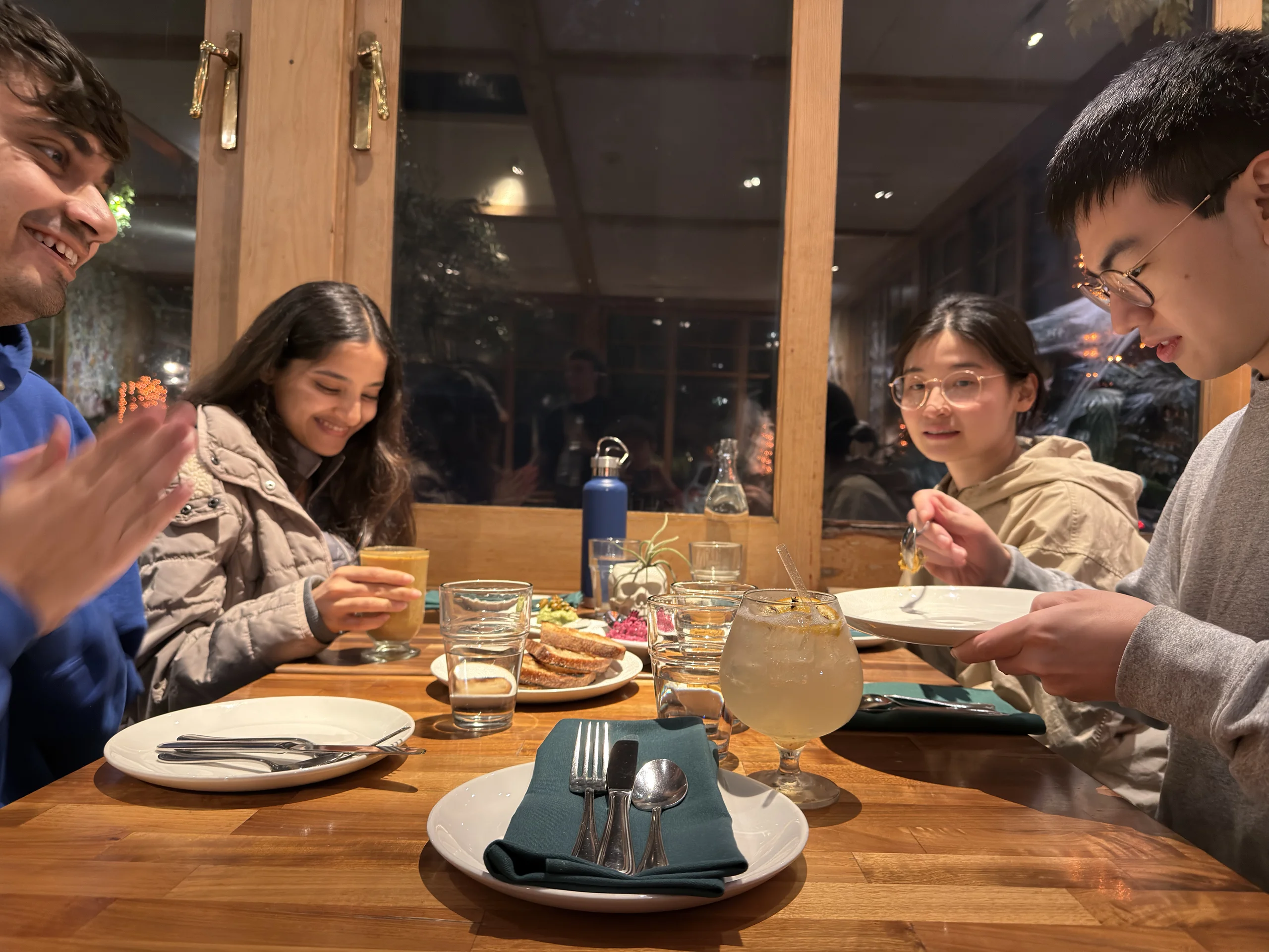 多人圍坐在木桌旁用餐,桌上有麵包、飲料與餐具,室內燈光溫暖