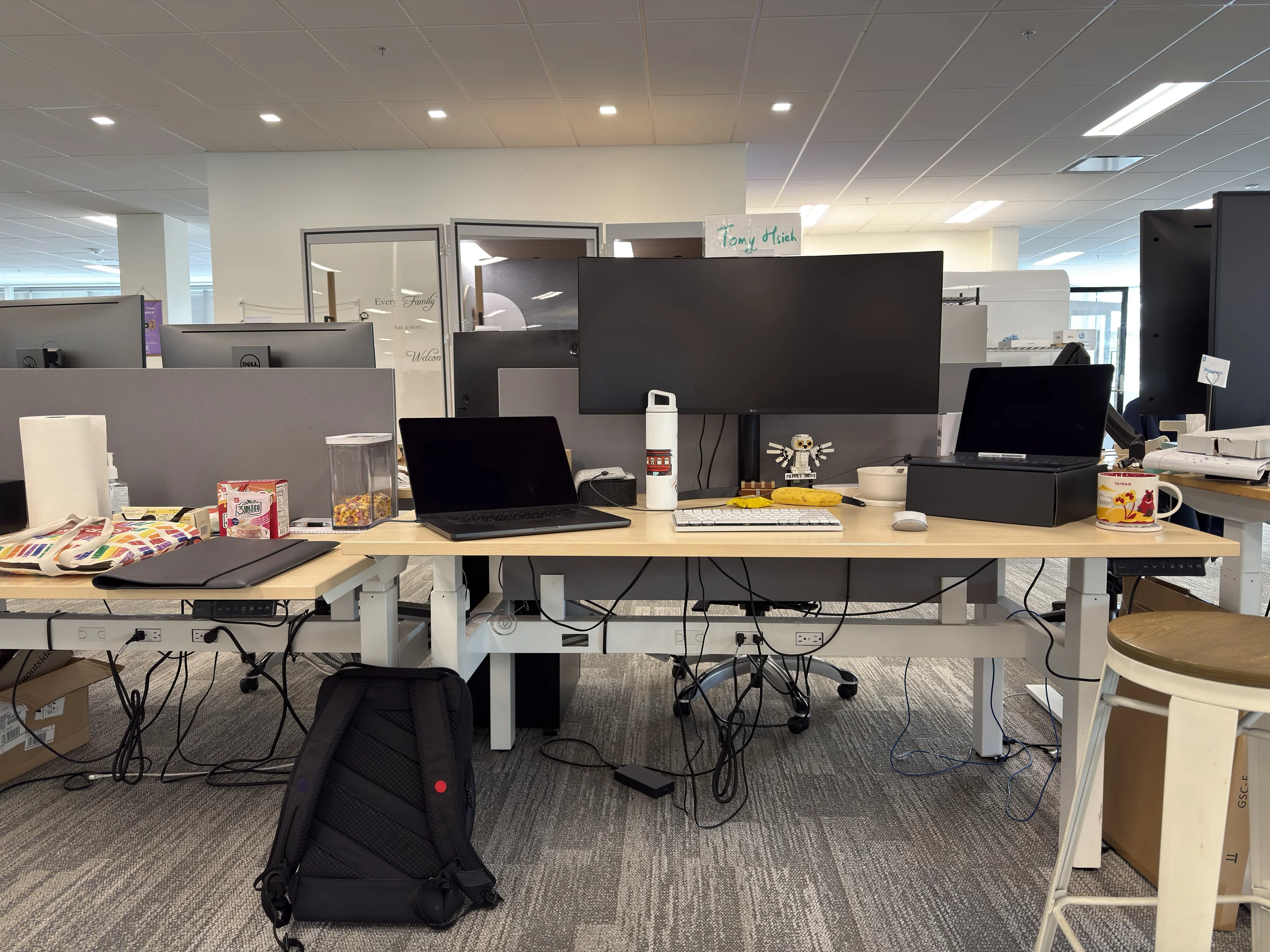 辦公室桌面擺放筆電、螢幕與零食，桌下放著黑色背包