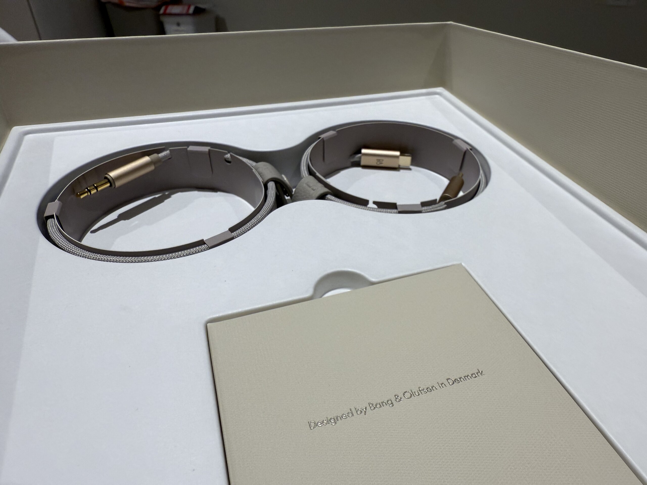 Beoplay H100 包裝內部視圖，展示兩個圓形隔層，內含音訊線材，搭配高級布質紋理，並附有一張印有『Designed by Bang & Olufsen in Denmark』字樣的小盒子。