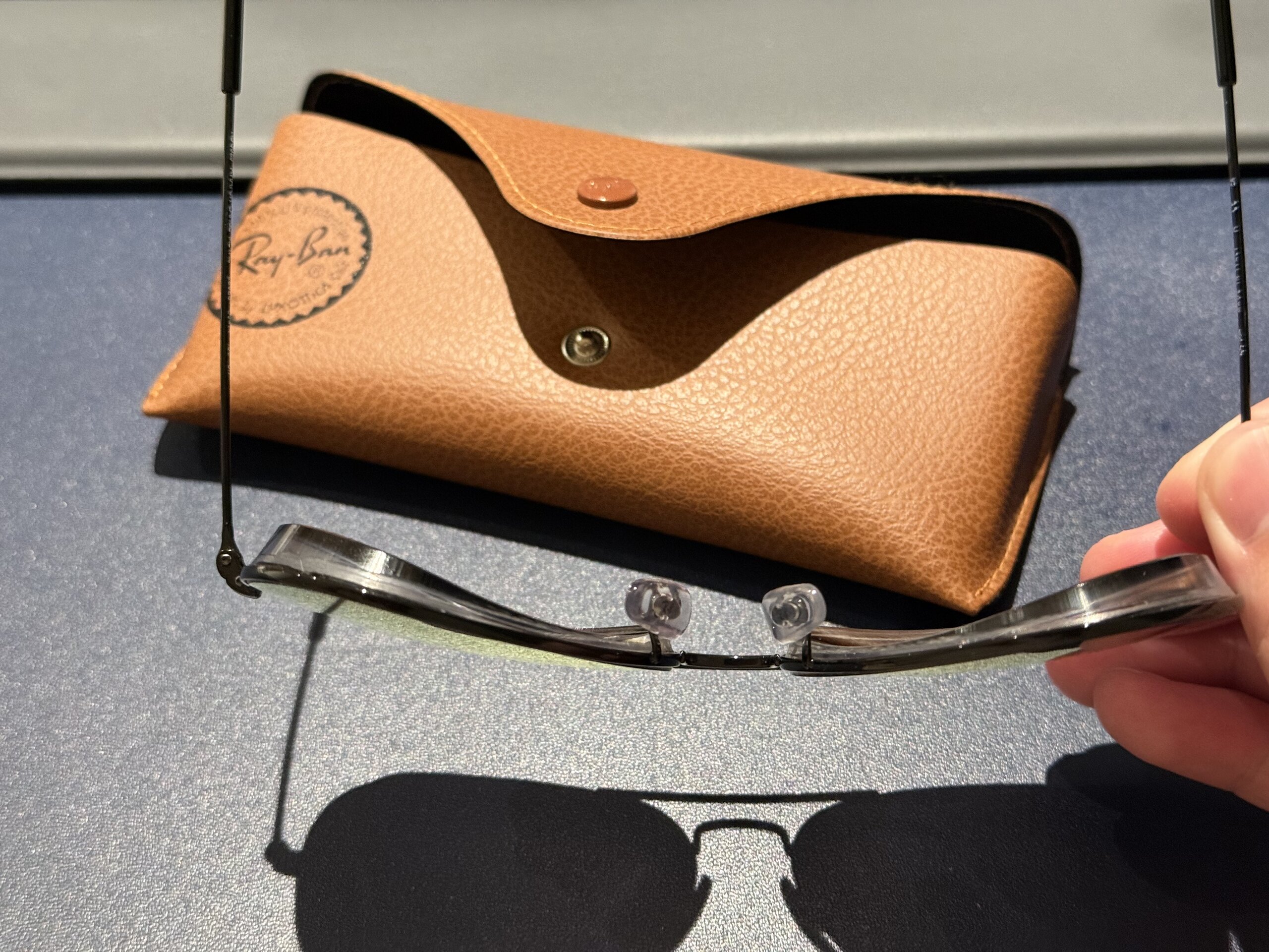 手持 Ray-Ban 墨鏡並展示鏡片厚度，背景有棕色皮革眼鏡盒