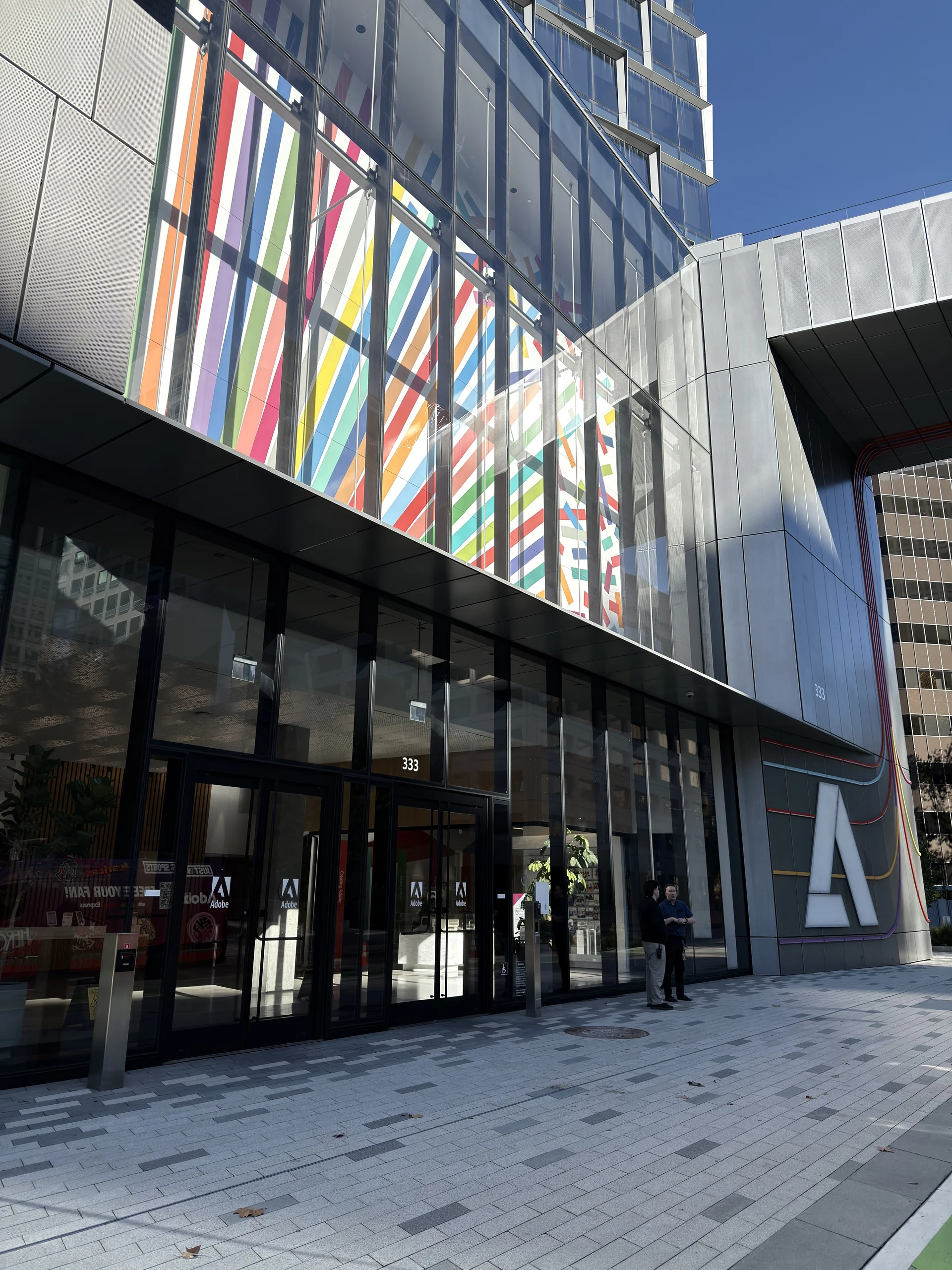 Adobe 總部大樓外觀，建築立面帶有彩色幾何裝飾，入口處可見 Adobe 標誌與行人，整體風格現代且具設計感。