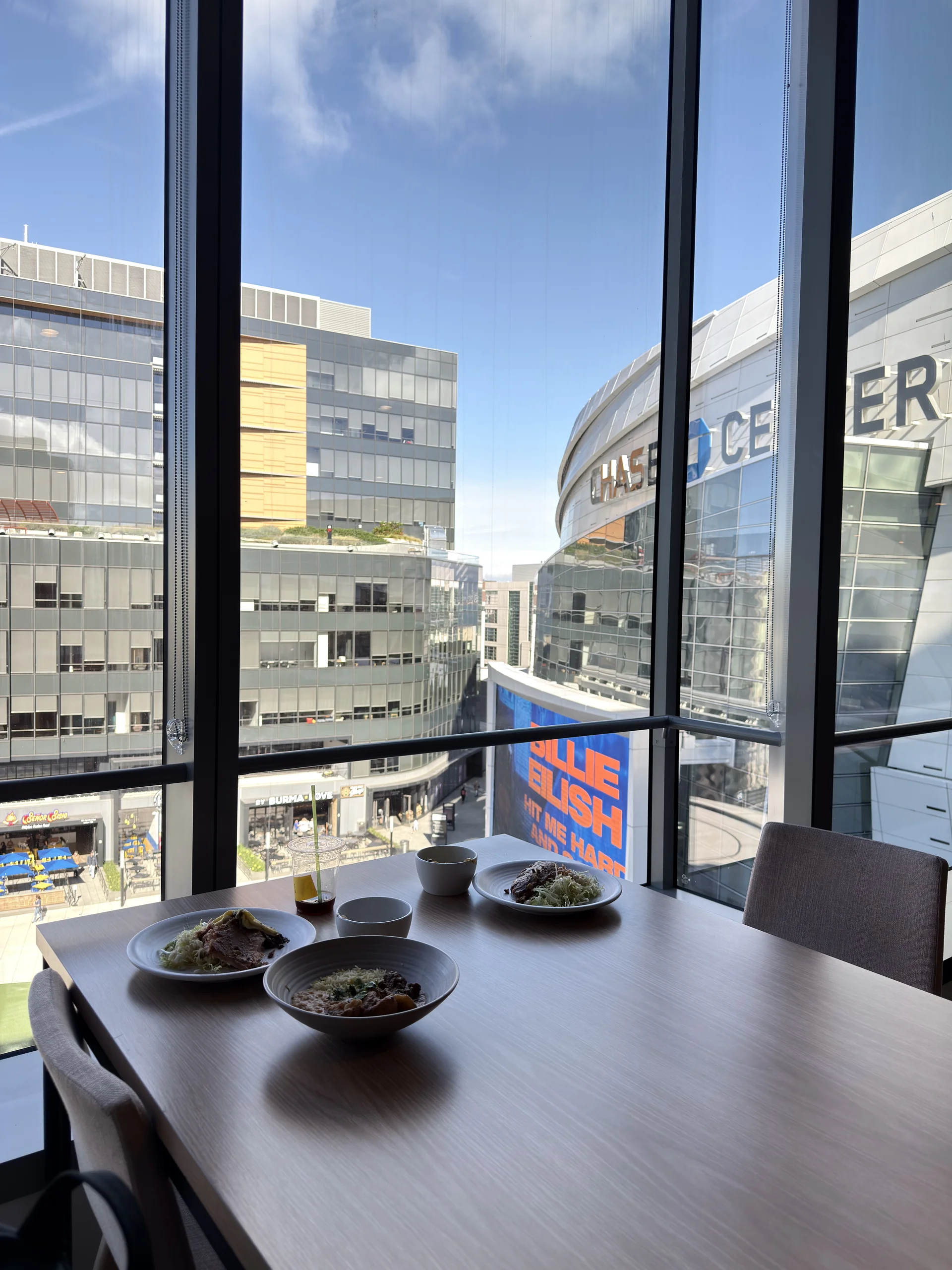 室內高樓餐廳的用餐桌景，桌上擺放簡單餐點，透過大片落地窗可見城市街景與大型建築外牆廣告，光線明亮、構圖乾淨。