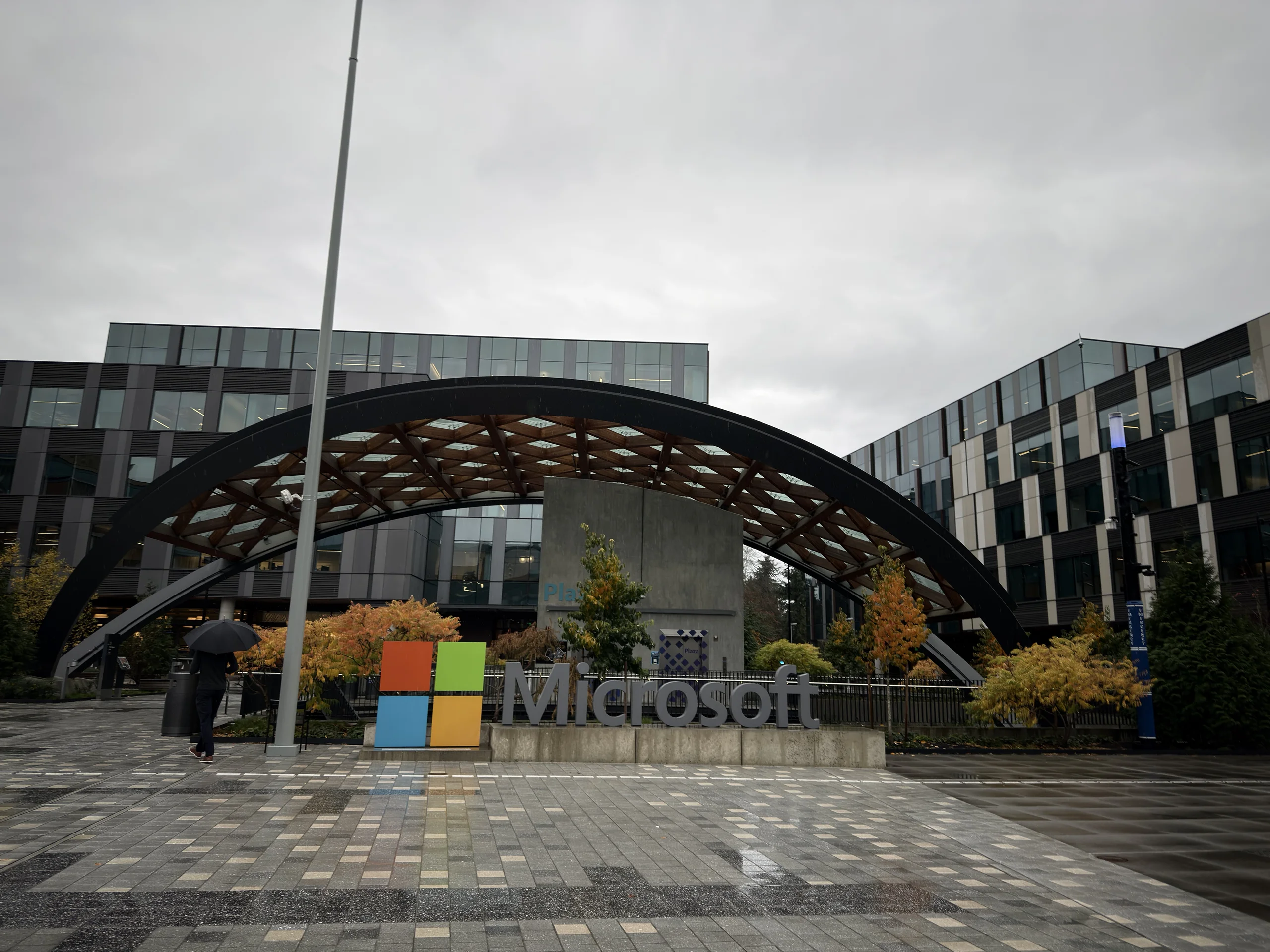 Microsoft 園區入口景觀，前景是 Microsoft 標誌，後方是現代感建築與拱形結構，天空陰雨，整體呈現企業總部的沉穩氛圍。