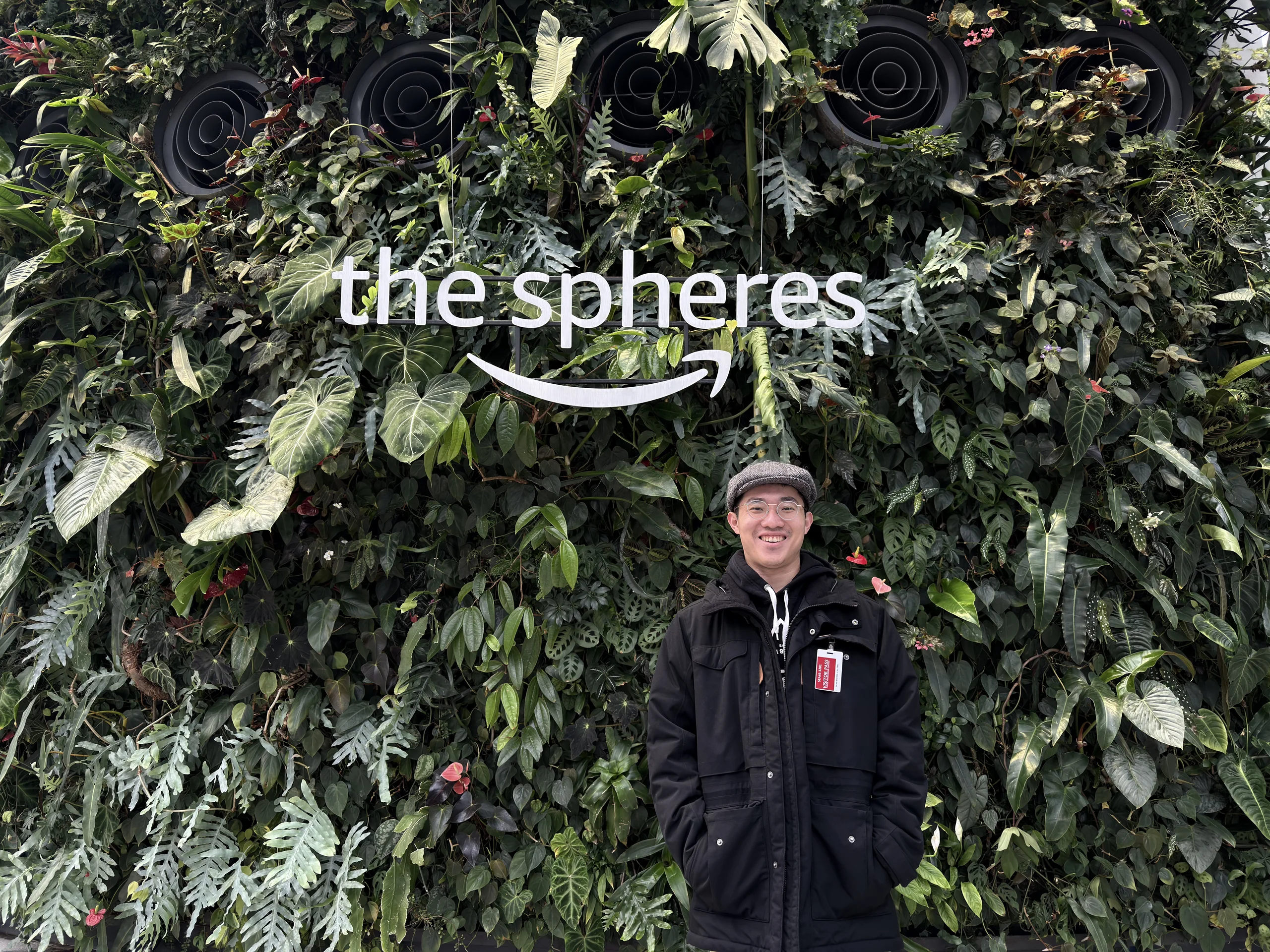 Tomy 站在 Amazon The Spheres 的植物牆前，牆面佈滿綠色植栽並寫有「the spheres」字樣，呈現科技園區與自然融合的設計風格。
